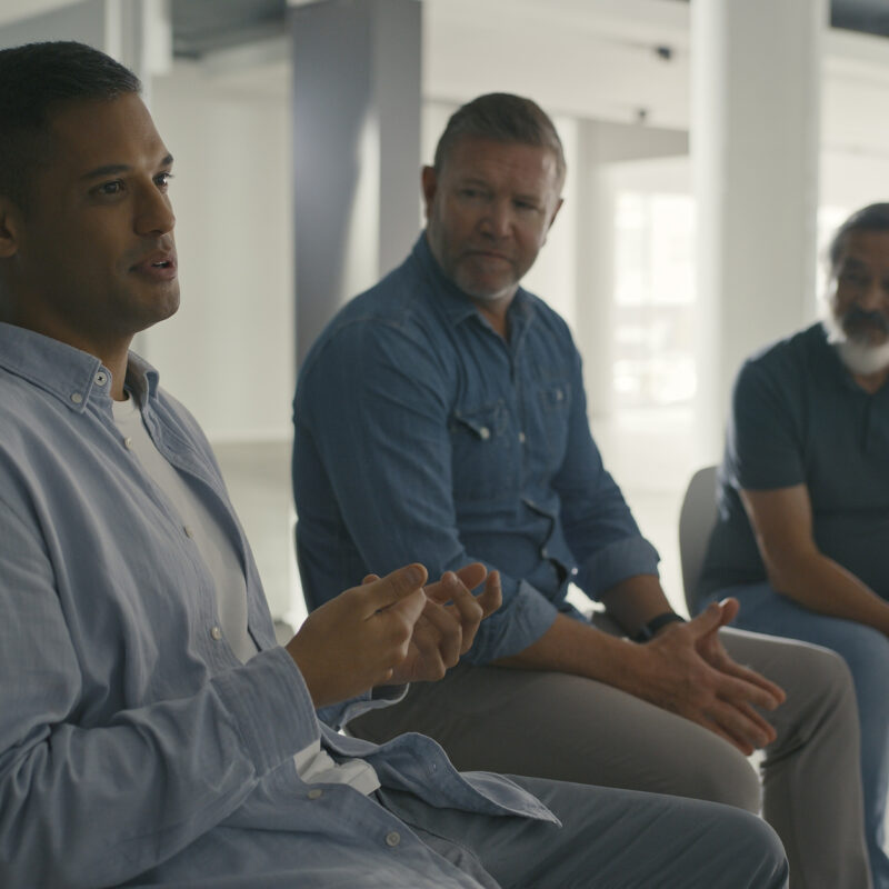Men in an AA support group meeting for alcohol abuse, talking while sitting in a circle during a therapy session. Male colleagues discussing feelings and emotions in a team psychology meeting
